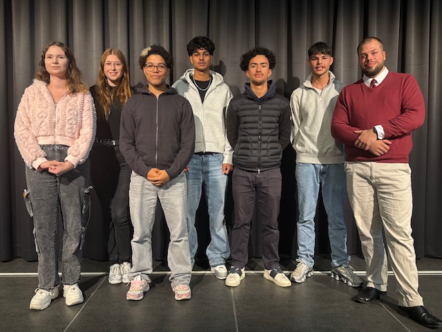 Sieben Personen stehen in zwei Reihen auf einer Bühne vor einem schwarzen Vorhang. Die vordere Reihe besteht aus fünf Personen, die hintere aus zwei Personen. Alle tragen Freizeit- oder Business-Kleidung, darunter Jeans, Sneakers, Pullover und eine Person mit rotem Pullover und weißem Hemd. Der Boden ist dunkel und die Beleuchtung kommt von vorne.